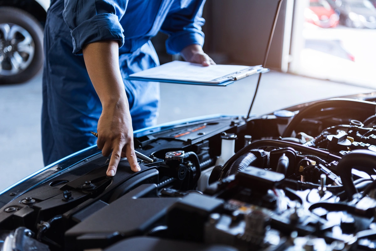 Technicien en tenue bleue effectuant une inspection du moteur d'un véhicule, avec une tablette de diagnostic à la main.
